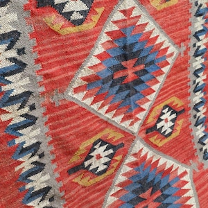 K&ouml;nnte beinhalten: Nahaufnahme eines lebendigen, handgewebten Teppichs mit geometrischem Muster. Der Teppich hat einen roten Hintergrund mit Rauten- und Zickzack-Designs in Blau, Wei&szlig;, Gelb und Schwarz. Die Textur des Teppichs ist sichtbar und zeigt die Handwerkskunst.