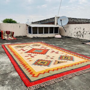 Pode incluir: Um tapete colorido tecido &agrave; m&atilde;o com um padr&atilde;o geom&eacute;trico em tons de vermelho, amarelo, verde e azul. O tapete tem uma borda de franjas e est&aacute; colocado sobre uma superf&iacute;cie de concreto.