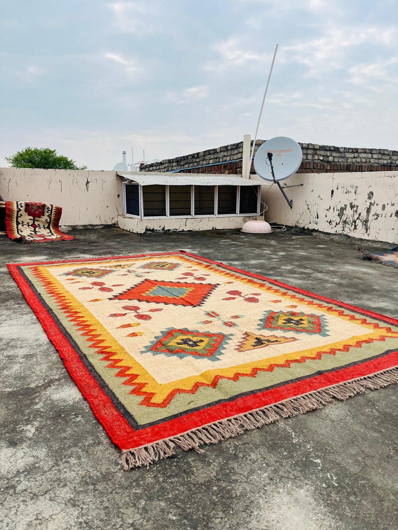 Pode incluir: Um tapete colorido tecido &agrave; m&atilde;o com um padr&atilde;o geom&eacute;trico em tons de vermelho, amarelo, verde e azul. O tapete tem uma borda de franjas e est&aacute; colocado sobre uma superf&iacute;cie de concreto.