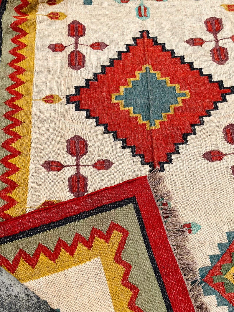 Pode incluir: Um tapete branco com um padr&atilde;o geom&eacute;trico em tons de vermelho, amarelo, verde e azul. O tapete tem uma borda de franjas.