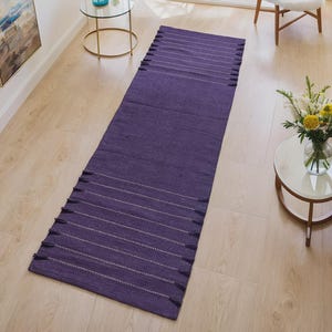 May include: A long, rectangular purple rug with white horizontal stripes and fringe on both ends. The rug is made of a woven material and is placed on a light-colored wooden floor. The rug is in a living room setting.