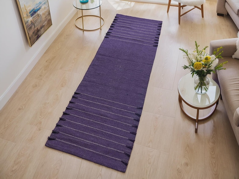 May include: A long, rectangular purple rug with white horizontal stripes and fringe on both ends. The rug is made of a woven material and is placed on a light-colored wooden floor. The rug is in a living room setting.