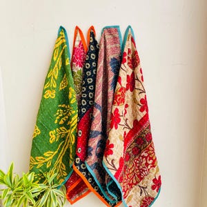 May include: Five colorful, patterned fabric pieces hanging against a white wall. The fabrics feature floral and geometric designs in shades of green, red, blue, and orange. Each piece has a contrasting border, adding to the visual appeal. The fabrics appear to be handmade.