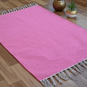 May include: A rectangular pink rug with a woven texture and fringed edges. The rug is displayed on a wooden floor, with decorative items in the background. The rug's dimensions are not visible.