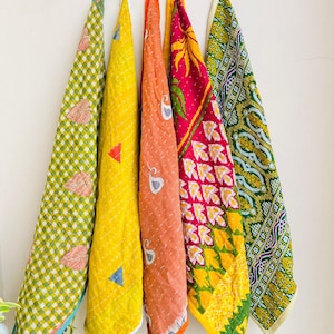 May include: Five vibrant, patterned textiles hanging against a white wall. The textiles display diverse designs, including geometric shapes, floral patterns, and a mix of colours like yellow, orange, pink, and green. The edges are stitched with contrasting thread.