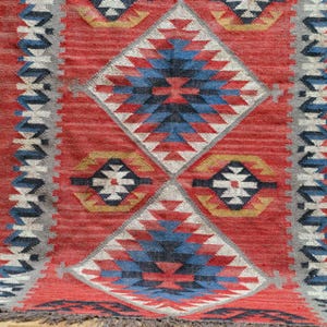 K&ouml;nnte beinhalten: Ein roter Teppich mit geometrischem Muster aus Rauten und Rechtecken. Der Teppich hat blaue, wei&szlig;e, gelbe und graue Akzente. Der Teppich hat einen Fransen am unteren Rand.