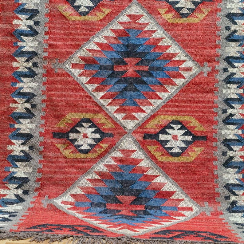 K&ouml;nnte beinhalten: Ein roter Teppich mit geometrischem Muster aus Rauten und Rechtecken. Der Teppich hat blaue, wei&szlig;e, gelbe und graue Akzente. Der Teppich hat einen Fransen am unteren Rand.
