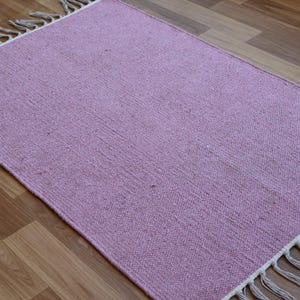 May include: A rectangular, woven rug in a soft lavender hue. The rug features a textured surface and is trimmed with a fringe detail in a neutral color. The rug is placed on a wooden floor.