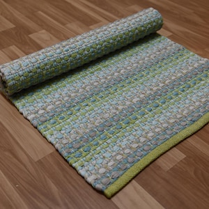 May include: A rolled-up, rectangular rug with a woven texture. The rug features a pattern of blue, green, and gray horizontal stripes. A green border frames the edge of the rug. The rug is partially rolled up, revealing the underside.