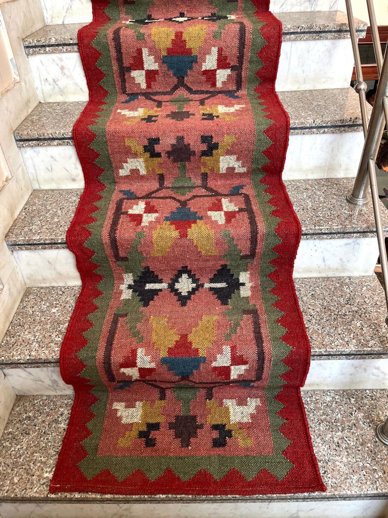 May include: A woven stair runner rug with a geometric pattern. The rug features a central design in shades of pink, red, green, yellow, and blue. The edges are trimmed with a red border. The rug is placed on a set of stairs.