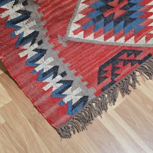 K&ouml;nnte beinhalten: Ein roter, blauer und grauer gewebter Teppich mit geometrischem Muster. Der Teppich hat ein Diamanten- und Pfeildesign mit einem Fransenrand. Der Teppich besteht aus einem strukturierten Material und liegt auf einem Holzboden.