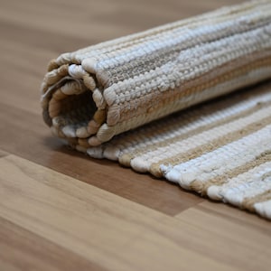 Op de afbeelding: Opgerolde, gestreepte vloerkleed in neutrale tinten beige, bruin en wit. Het vloerkleed is gemaakt van geweven materiaal en heeft een textuur. De strepen lopen in de lengte en het vloerkleed is gedeeltelijk uitgerold.