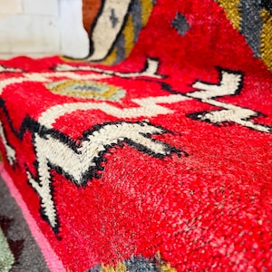 Puede incluir: Primer plano de una alfombra tejida roja, blanca, negra y dorada con un patr&oacute;n geom&eacute;trico. La alfombra est&aacute; hecha de lana y tiene una superficie suave y texturizada.