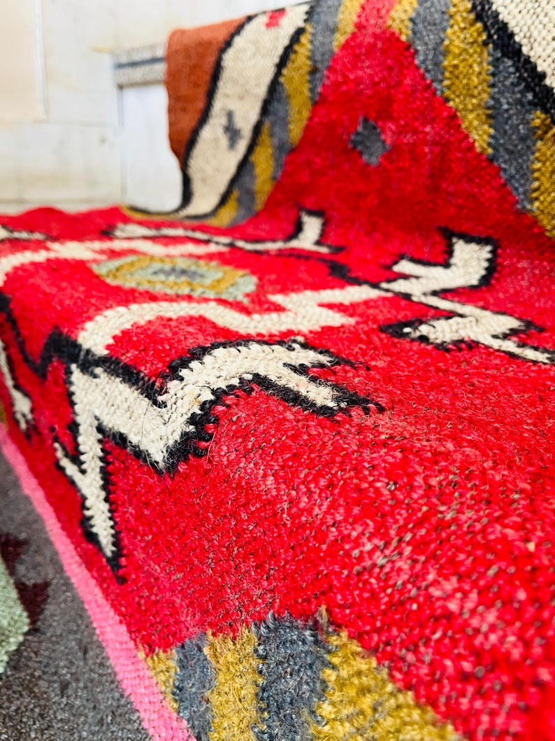 Puede incluir: Primer plano de una alfombra tejida roja, blanca, negra y dorada con un patr&oacute;n geom&eacute;trico. La alfombra est&aacute; hecha de lana y tiene una superficie suave y texturizada.