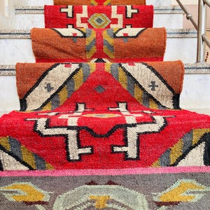 Puede incluir: Un conjunto de alfombras tejidas de colores con patrones geom&eacute;tricos en tonos rojos, naranjas, amarillos y grises. Las alfombras est&aacute;n dispuestas en un conjunto de escaleras, creando una exhibici&oacute;n visualmente atractiva.