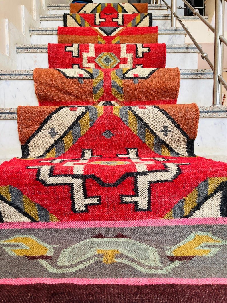 Puede incluir: Un conjunto de alfombras tejidas de colores con patrones geom&eacute;tricos en tonos rojos, naranjas, amarillos y grises. Las alfombras est&aacute;n dispuestas en un conjunto de escaleras, creando una exhibici&oacute;n visualmente atractiva.