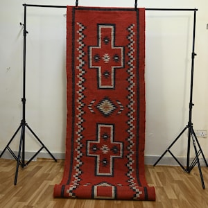 May include: A red and black woven runner rug with a geometric cross and diamond pattern. The rug is displayed vertically, showcasing its design and texture. The rug is made of a thick material and has a rolled-up bottom.
