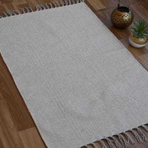 May include: A rectangular, light gray rug with a woven texture and fringed edges. The rug is displayed on a wooden floor, with decorative items including a gold lantern and a small potted plant.