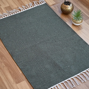 May include: A rectangular, dark green rug with white fringe trim. The rug is made of woven material and is placed on a wooden floor. Decorative items are in the background.