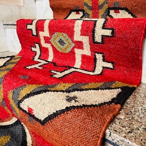 Puede incluir: Una alfombra roja, marr&oacute;n y blanca con dise&ntilde;os geom&eacute;tricos. La alfombra est&aacute; hecha de tejido tejido y tiene un borde con flecos.