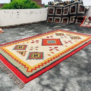Pode incluir: Um tapete colorido com um padr&atilde;o geom&eacute;trico em tons de vermelho, amarelo, azul e verde. O tapete tem uma borda com franjas e est&aacute; colocado sobre uma superf&iacute;cie de concreto.