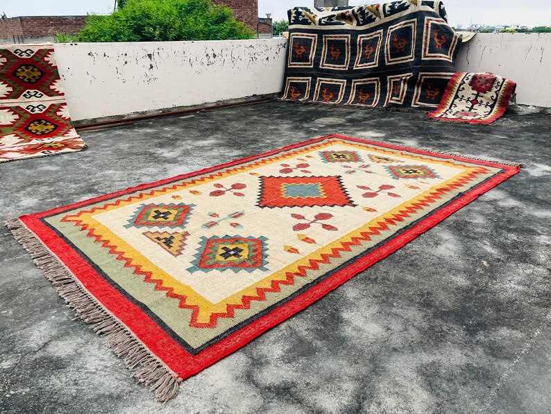 Pode incluir: Um tapete colorido com um padr&atilde;o geom&eacute;trico em tons de vermelho, amarelo, azul e verde. O tapete tem uma borda com franjas e est&aacute; colocado sobre uma superf&iacute;cie de concreto.