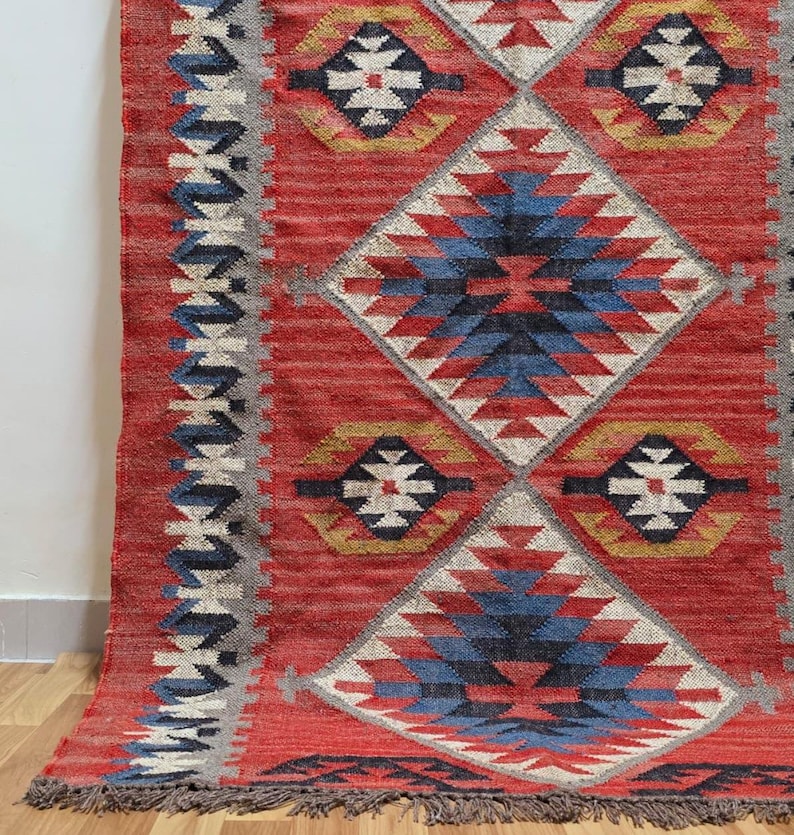 K&ouml;nnte beinhalten: Ein roter gewebter Teppich mit einem geometrischen Rautenmuster. Der Teppich hat Rauten in Rot, Blau, Wei&szlig; und Gelb. Die Kanten haben einen Fransen und einen Rand mit einem sich wiederholenden Muster aus Blau, Wei&szlig; und Grau.