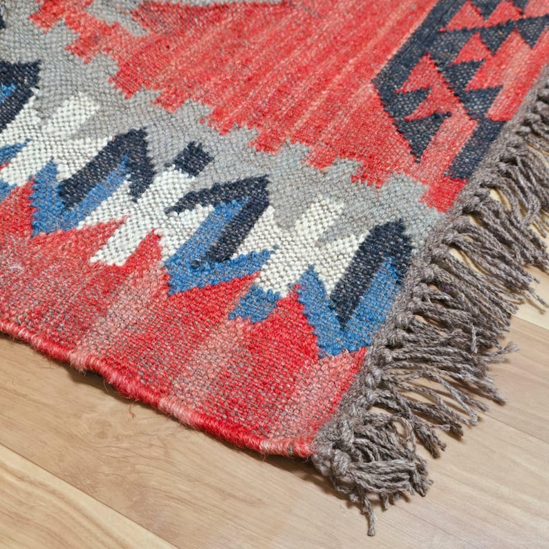 May include: Close-up of a woven rug with a geometric pattern. The rug features a combination of red, gray, blue, and white colors. The rug has a fringed edge and is placed on a wooden floor.