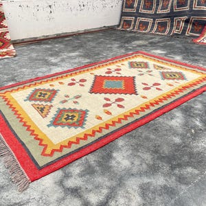 Pode incluir: Um tapete colorido com um padr&atilde;o geom&eacute;trico em tons de vermelho, amarelo, azul e verde. O tapete tem um fundo branco e uma borda com franjas.