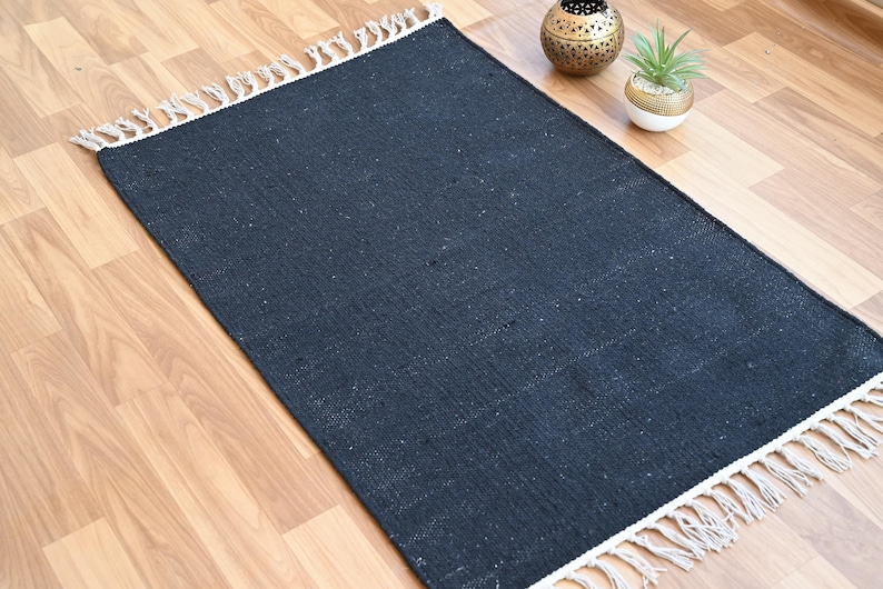 May include: A rectangular, dark blue rug with a woven texture and white fringe trim. The rug is placed on a light wood floor. Decorative items, including a gold-colored metal lantern and a small potted plant, are visible in the background.