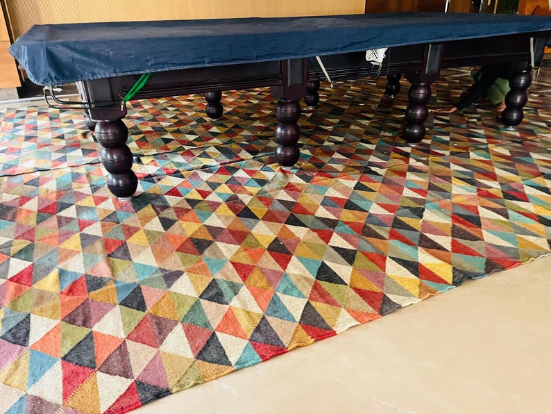 May include: A dark blue pool table cover with a patterned rug underneath. The table has six legs and is made of dark wood.