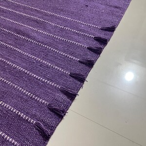 May include: A purple woven rug with white horizontal stripes and tassels on the edge. The rug is made of a textured material and is partially visible, with a white floor in the background.