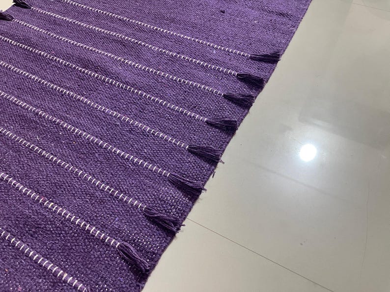 May include: A purple woven rug with white horizontal stripes and tassels on the edge. The rug is made of a textured material and is partially visible, with a white floor in the background.