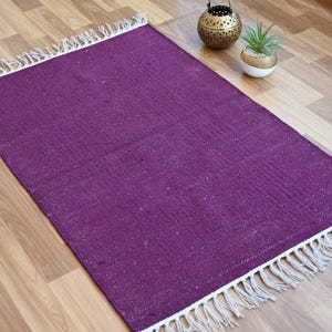 May include: A rectangular, purple rug with white fringe along the short ends. The rug has a woven texture and is placed on a light brown wooden floor. Decorative items are in the background.