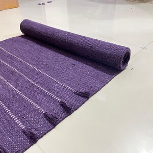 May include: A rolled-up purple rug with white striped accents and tassels. The rug is made of woven material and is partially unrolled, displaying its texture and design. The rug is on a white tiled floor.