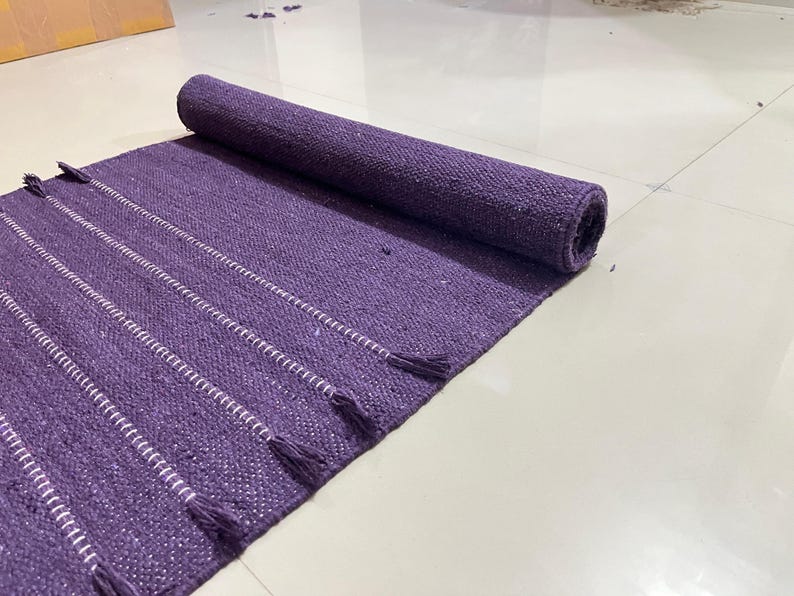 May include: A rolled-up purple rug with white striped accents and tassels. The rug is made of woven material and is partially unrolled, displaying its texture and design. The rug is on a white tiled floor.