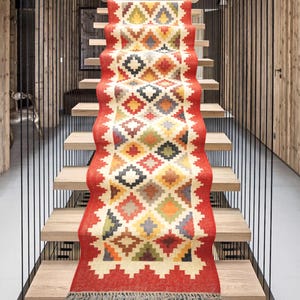 以下が含まれることがあります： 赤、オレンジ、黄色、緑、青、黒の色合いのカラフルな幾何学模様の敷物ランナー。ランナーは木製の階段に敷かれています。