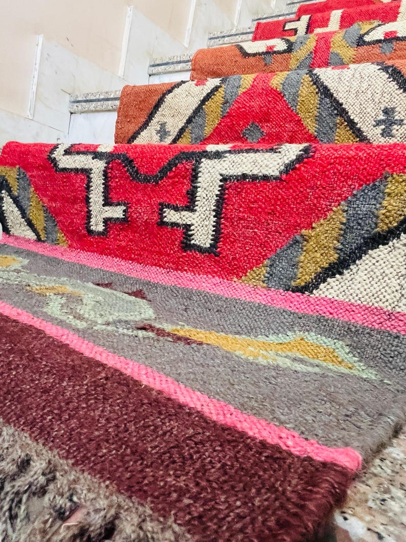 Puede incluir: Un conjunto de alfombras tejidas de colores con patrones geom&eacute;tricos en tonos rojos, rosas, marrones, grises y amarillos. Las alfombras est&aacute;n superpuestas en un tramo de escaleras.