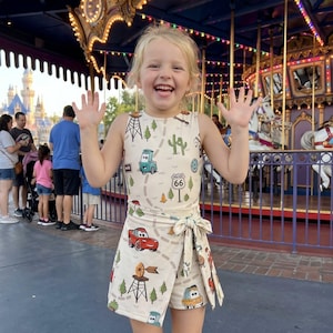 May include: A young child wearing a cream-colored romper with a Route 66 and car-themed print. The outfit includes a sleeveless top and wrap-around shorts. The child is smiling and waving in front of a carousel.