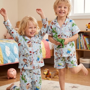 Puede incluir: Dos ni&ntilde;os con pijamas azul claro con estampado de Toy Story. Los pijamas tienen un patr&oacute;n de rayas y presentan personajes de la pel&iacute;cula. Un ni&ntilde;o est&aacute; arrodillado, mientras que el otro sostiene un juguete de dinosaurio verde. Una hucha rosa y un perro de juguete est&aacute;n en el fondo.