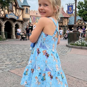 Pu&ograve; includere: Un vestito azzurro chiaro con personaggi del film Frozen. L'abito ha una gonna a balze e spalline incrociate. La bambina tiene in mano una bambola di Elsa.