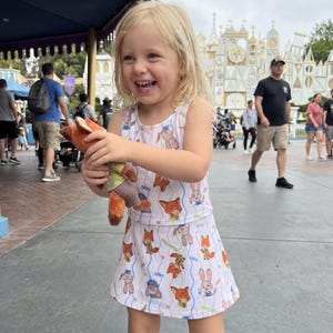 May include: A young child wearing a white dress with a repeating pattern of cartoon foxes and bunnies, holding a plush fox toy. The child is smiling and appears to be in a theme park setting.
