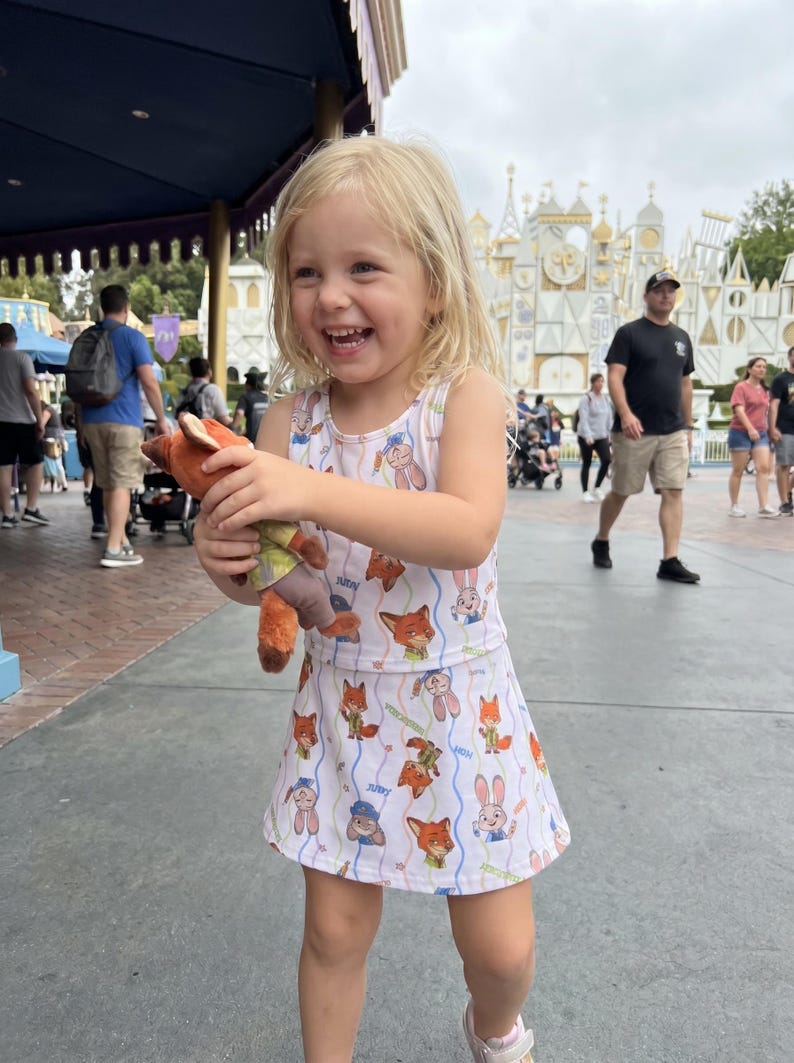 May include: A young child wearing a white dress with a repeating pattern of cartoon foxes and bunnies, holding a plush fox toy. The child is smiling and appears to be in a theme park setting.