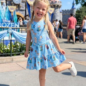 Pu&ograve; includere: Un vestito azzurro senza maniche con un motivo di personaggi del film "Frozen". L'abito ha una gonna a balze e viene indossato con scarpe da ginnastica bianche. Lo sfondo include un castello e le parole "Disney Frozen".