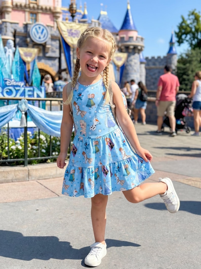 Pu&ograve; includere: Un vestito azzurro senza maniche con un motivo di personaggi del film "Frozen". L'abito ha una gonna a balze e viene indossato con scarpe da ginnastica bianche. Lo sfondo include un castello e le parole "Disney Frozen".