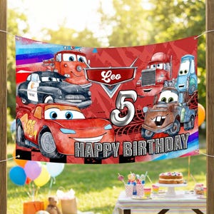 May include: A red banner featuring cartoon cars with the text "Leo" and "5 HAPPY BIRTHDAY". The banner is displayed outdoors with a party scene in the background, including balloons and a table with treats.