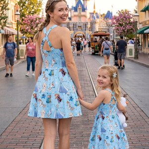 Pu&ograve; includere: Una madre e una bambina indossano abiti azzurri abbinati con una gonna a balze e schiena incrociata. Gli abiti presentano una stampa di personaggi del film Frozen. Stanno camminando per una strada lastricata di mattoni di fronte a un castello.