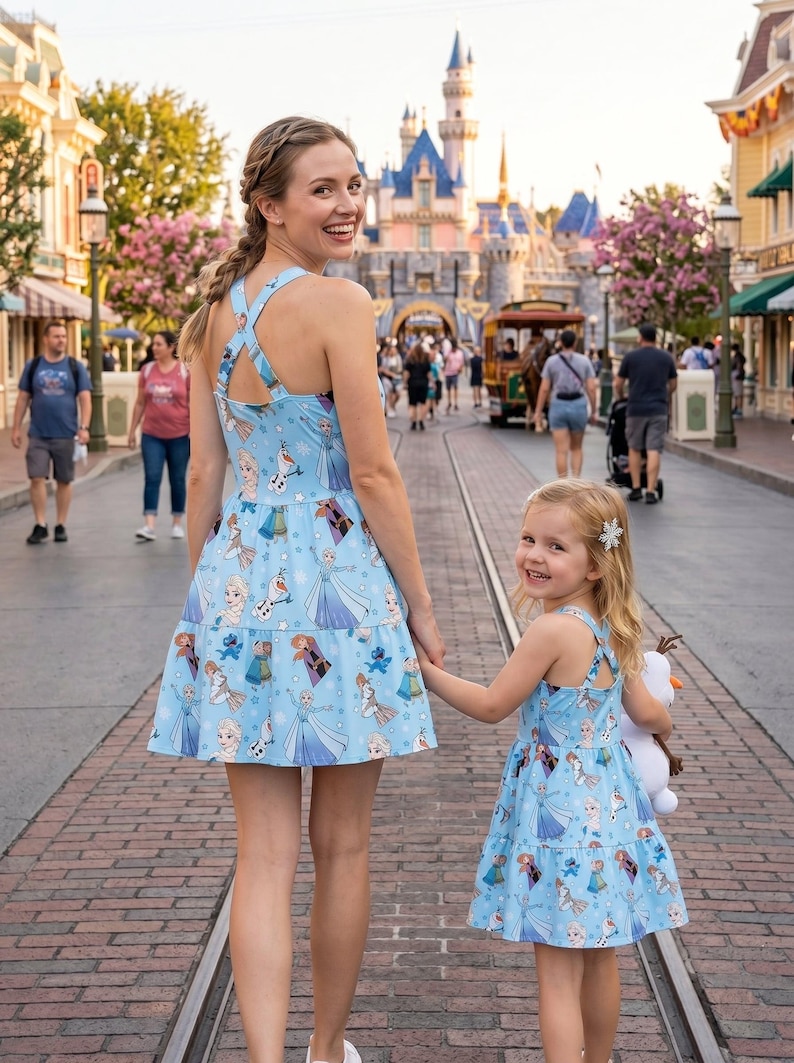 Pu&ograve; includere: Una madre e una bambina indossano abiti azzurri abbinati con una gonna a balze e schiena incrociata. Gli abiti presentano una stampa di personaggi del film Frozen. Stanno camminando per una strada lastricata di mattoni di fronte a un castello.