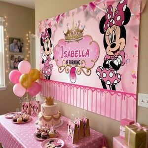May include: A pink Minnie Mouse-themed birthday party setup. A banner with Minnie Mouse and the name "Isabella" is displayed. The table is set with a cake, cupcakes, plates, gift bags, balloons, and wrapped gifts.