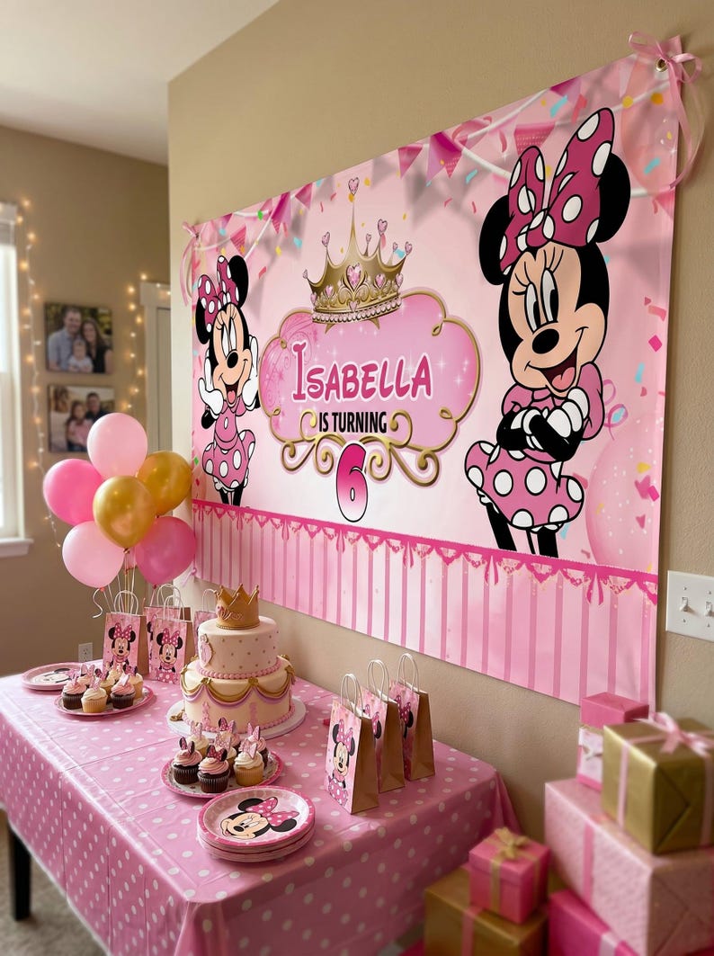 May include: A pink Minnie Mouse-themed birthday party setup. A banner with Minnie Mouse and the name "Isabella" is displayed. The table is set with a cake, cupcakes, plates, gift bags, balloons, and wrapped gifts.